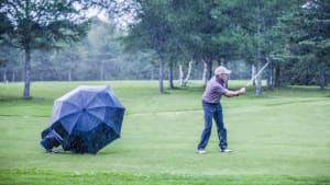 A person is playing golf in the rain on a green course, with a collapsed black umbrella nearby. Trees are visible in the background.